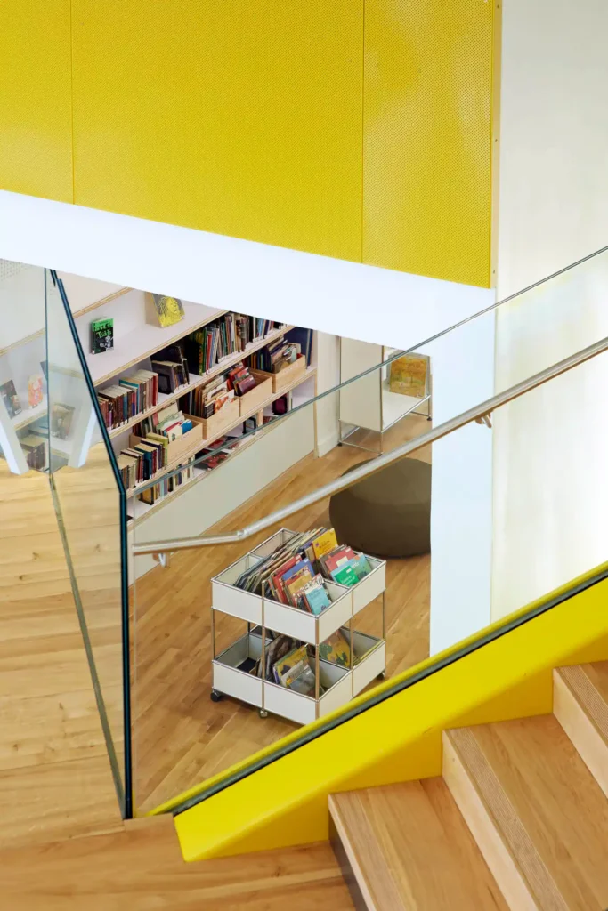 Vue intérieure moderne de la médiathèque B612 avec rayonnages de livres jeunesse et escalier aux finitions bois et jaune.